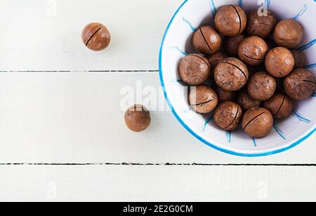 Organic Macadamia nuts (Macadamia integrifolia) in shell on wooden white table. Top view, overhead, copy space Stock Photo