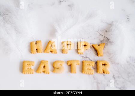 Lettering from biscuits in form of letters and white fluffy plumelets on marble background. Happy Easter background, creative greeting. Stock Photo