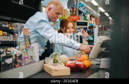 Senior manager training a new female employee at checkout counter. New cashier getting some help from store manager. Stock Photo