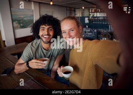 Portrait of cheerful young couple in modern cafe relaxing holding coffee mug and taking selfie Stock Photo