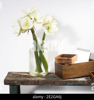 Bouquet of three white lilies in a tall glass vase on a beige vintage bench against a gray wall. Wodden boxes different size with scetchbook. Copy spa Stock Photo