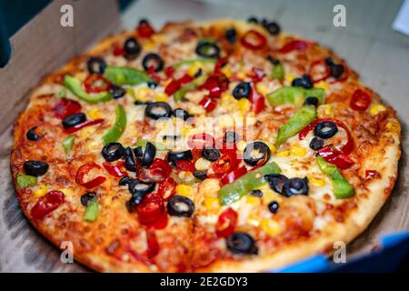 Selective focus of a vegetarian pan pizza topped with black olives, green bell peppers, red paprika, and golden corn in a takeout box Stock Photo