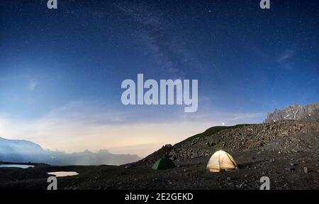 Magic scenery at night with blue sky and shining stars, beautiful mountains area with snow and lakes. Gorgeous mountain ridge with high rocky peaks, two colorful tourist tents. Panoramic view Stock Photo