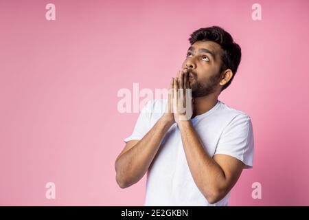 Closeup of young indian male in white t shirt, praying with palms clasped together, asking the God for good luck, success, health, on pink background Stock Photo