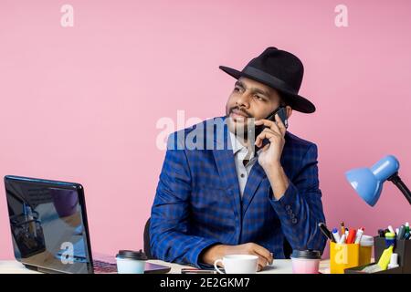 Joyful stylish indian entrepreneur wearing formal wear, black hat, talking on mobile phone, smiling broadly, looking to side, sitting at desk in offic Stock Photo