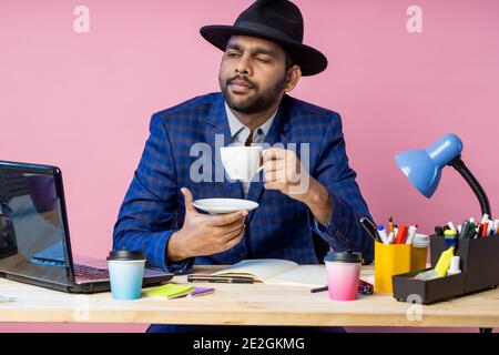 young unshaven indian businessman in formal blue checkered suit, black hat , sitting at workplace, thinking about something, looking aside, enjoying a Stock Photo