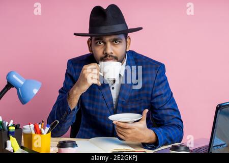 businessman in formal checkered suit, black hat , sitting at workplace, thinking about something, looking aside, enjoying aromatic coffee. Business, l Stock Photo