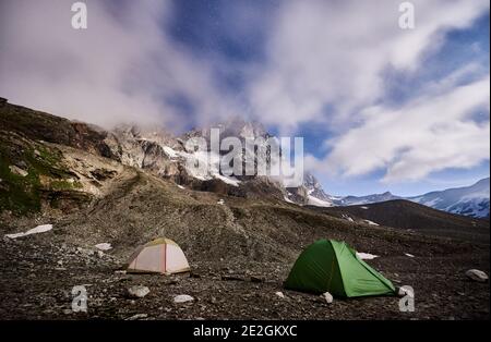 Panoramic view of night camping in night mountains. Scenery of tourist tents on rocky hill and mountain peak Matterhorn under beautiful misty sky with stars. Concept of travelling, hiking and camping. Stock Photo