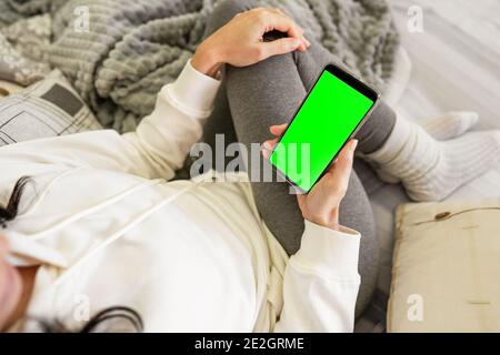 Top view of unrecognizable young woman on sofa holding smartphone with green screen blank display for copy space. Caucasian female beautiful housewife Stock Photo