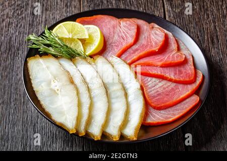 Smoked fish appetizer plate: cold smoked halibut and cold smoked tuna slices served on a black plate with rosemary sprigs and lemon wedges on a dark w Stock Photo