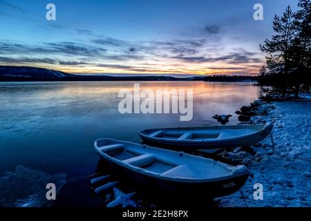 Two wooden rowing boats / rowboats on the shore of frozen lake at sunset in winter, Laponia, Lapland, Sweden Stock Photo