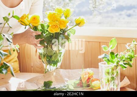 Woman arranging fresh spring yellow flowers in a glass vase on a wooden table near the window. Yellow roses. Natural light. Soft focus. Stock Photo