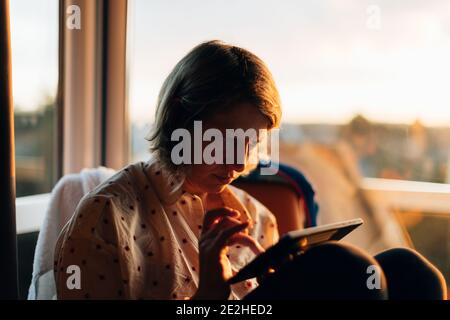 a woman sitting indoors nearby a big window at sunset using a tablet. Wireless technology concept Stock Photo