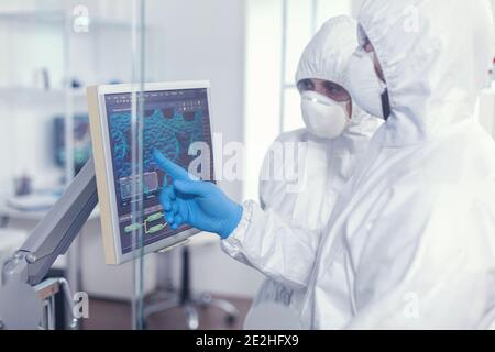Chemists in protection suit discussing about changes in vaccine composition in equipped lab pointing desktop. Doctors examining virus evolution using high tech researching diagnosis against covid19 Stock Photo
