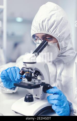 Woman scientis in coverall doing coronavirus investigation looking through microscope. Chemist researcher during global pandemic with covid-19 checking sample in biochemistry lab Stock Photo