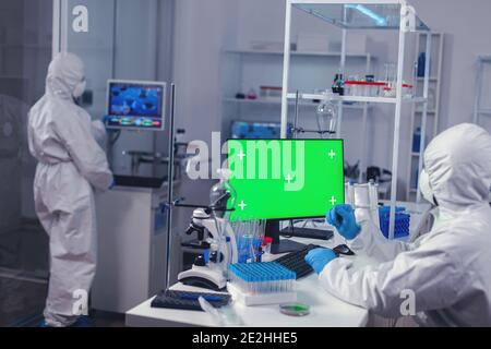 Scientist typing on keyboard works with computer with green screen dressed in ppe. Team of microbiologists doing vaccine research writing on device with chroma key, isolated, mockup display. Stock Photo