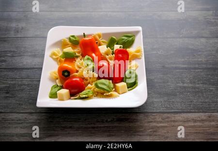 Fresh ingredients for a healthy salad with noodles, tomatoes, bell pepper, cheese cubes and basil on a white square plate. Dark wooden background. Stock Photo