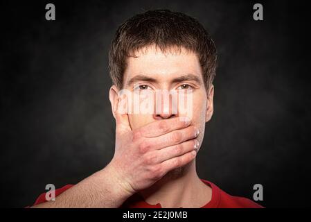 A man covers his mouth with his hand on a dark background Stock Photo