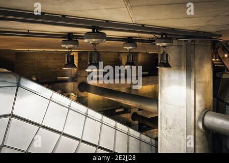 Industrial looking architecture, metal lamps shine over tubes, concrete walls and pillars in modern train station. Blurred people going down the escal Stock Photo