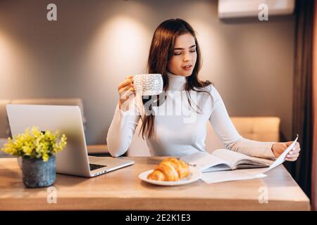 Freelance student young female using laptop while sitting at home. Young woman sitting in kitchen and working online on laptop. Pretty female drinking Stock Photo