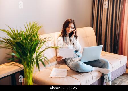 Freelance student young female using laptop while sitting at home. Young woman sitting on sofa and working online on laptop. Pretty female talking on Stock Photo