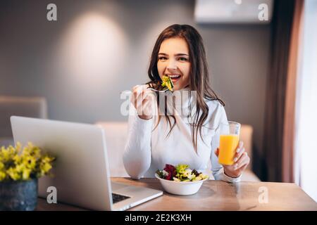 Smiling woman relaxing on her laptop and posing while drinking a glass of orange juice in her kitchen Stock Photo