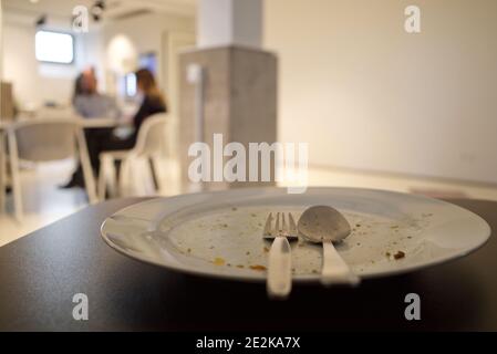 Empty and dirty white dish with stainless spoon and fork, and on black table after finish meal, and background of blur kitchen, office's canteen. Stock Photo