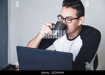Caucasian man drinking tea from a black mug while working from home and sitting in front of his laptop Stock Photo
