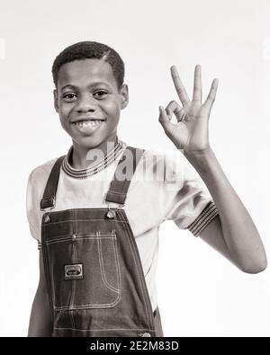 1970s SMILING AFRICAN-AMERICAN PRE-TEEN BOY WEARING BIB OVERALLS MAKING OKAY SIGN WITH HAND LOOKING AT CAMERA - b25650 HAR001 HARS MALES OK TEENAGE BOY DENIM GESTURING B&W EYE CONTACT DREAMS HAPPINESS CHEERFUL AFRICAN-AMERICANS AFRICAN-AMERICAN OKAY BLACK ETHNICITY GESTURES SMILES CONCEPTUAL JOYFUL BIB FINE INFORMAL JUVENILES OKEY-DOKEY PRE-TEEN PRE-TEEN BOY TWILL BLACK AND WHITE CASUAL HAR001 OLD FASHIONED AFRICAN AMERICANS Stock Photo