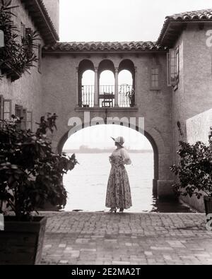 1920s ELEGANT WOMAN STANDING IN SPANISH TILE ROOF STYLE BUILDING GARDEN PATIO ARCHED WALKWAY OVER WATER FLORIDA USA - g1499 HAR001 HARS ABSTRACT RICH LIFESTYLE ARCHITECTURE FEMALES PATIO HOME LIFE LUXURY COPY SPACE FULL-LENGTH LADIES PERSONS ROOF B&W TIME OFF STRUCTURE STYLES TILE TRIP GETAWAY EXTERIOR ARCHED HOLIDAYS MOOD UPSCALE AFFLUENT STYLISH WALKWAY FASHIONS VACATIONS WELL-TO-DO YOUNG ADULT WOMAN ATMOSPHERE BLACK AND WHITE CAUCASIAN ETHNICITY HAR001 OLD FASHIONED Stock Photo