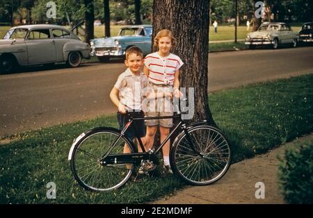 1950s GIRL AND BOY STANDING WITH BICYCLE LOOKING AT CAMERA ON SIDEWALK PARKED CARS ON STREET BEHIND - kb27433 KNA001 HARS STYLE PLEASED FAMILIES JOY FEMALES BROTHERS BIKING HEALTHINESS HOME LIFE COPY SPACE FRIENDSHIP HALF-LENGTH PHYSICAL FITNESS SNAPSHOT MALES SIBLINGS BICYCLES SISTERS TRANSPORTATION PARKED BIKES SUMMERTIME EYE CONTACT HAPPINESS WELLNESS AND SIBLING SMILES JOYFUL JUVENILES SEASON TOGETHERNESS CAUCASIAN ETHNICITY OLD FASHIONED Stock Photo