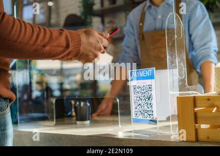 Asian man customer using mobile scan QR or NFC payment on smart phone paying for her coffee drink in coffee shop and plexi screen and Barista wearing Stock Photo