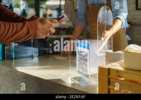 Asian man customer using mobile scan QR or NFC payment on smart phone paying for her coffee drink in coffee shop and plexi screen and Barista wearing Stock Photo