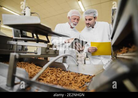Two business man in sterile clothes standing in food factory in front of the production line and looking and tablet. Checking the quality of products Stock Photo