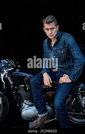 Handsome man wearing double denim sitting on motorcycle with black background . Portrait of handsome young man on vintage cafe racer motorcycle . Stock Photo
