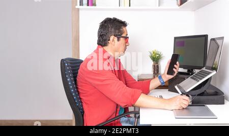 Man working at home with a computer and graphic tablet checks his smartphone. Latino man working at home Stock Photo