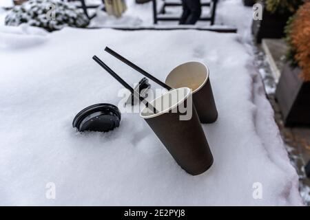 Two coffee cups and straws stick out in the snow on the terrace next to the street cafe