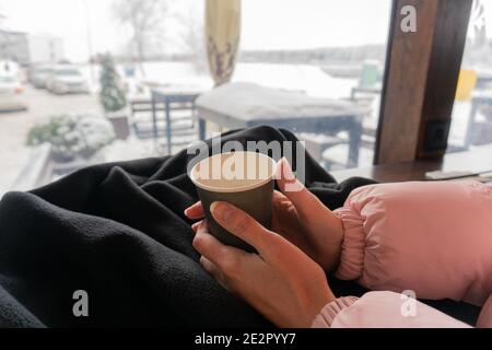 A paper cup of coffee in the hands of a girl in a cafe behind a bar with a view of a winter street