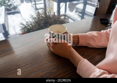 A girl in a pink sweater holds a paper cup with two hands with coffee on a winter day