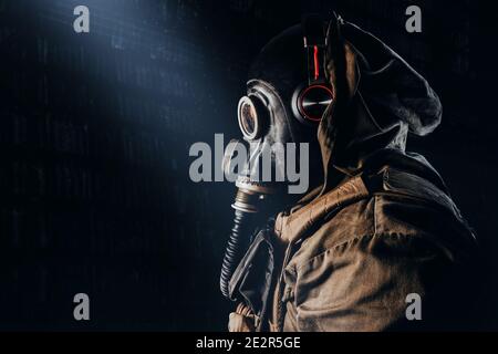 Photo of a stalker soldier in soviet rubber gas mask with  hose, wearing headphones and standing profile view on dark underground backdrop. Stock Photo