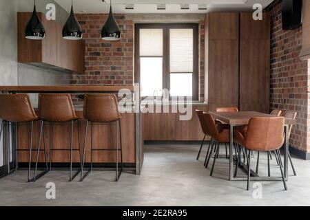 Contemporary interior design of light spacious dinning room including brown wooden furniture with bar stools at counter and soft comfortable chairs at Stock Photo