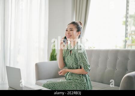 Happy young woman freelancer talking on mobile phone and using laptop, working from home during isolation, copy space. Stock Photo