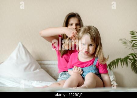 cute sister teen combing hair baby in bed.Cute girls are laughing and smiling. Morning treatments, hair care, care. Family love. Cozy bright bedroom. Stock Photo
