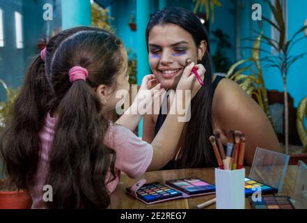 Girl with a brush in her hand putting makeup on a young woman. On a table there are three open makeup cases with various colors and various brushes Stock Photo