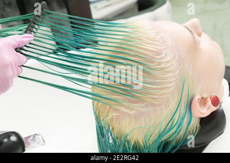 Professional hairstylist in protective glove holds of hair in hand and brushes long green and bleached hair of client, while washing head in shower Stock Photo