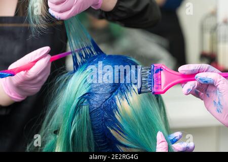 Two professional hairdressers in protective glove using pink brush while applying blue paint to female with emerald hair color, process of dyeing hair Stock Photo