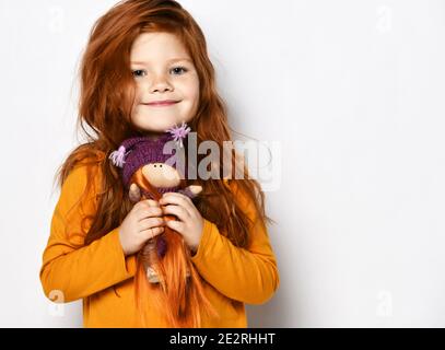 Little five-year-old red-haired kid girl in an orange sweatshirt smiles happily holding small redhair doll in her hands Stock Photo