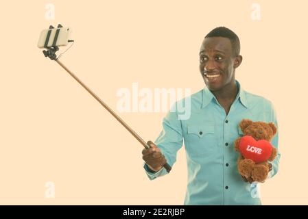 Young happy black African man smiling and holding teddy bear with heart and love sign while taking selfie picture with mobile phone on selfie stick Stock Photo