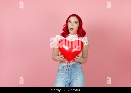 Close-up a surprised, shocked young woman with red hair holds a large flying red heart-shaped balloon isolated on a pink background Stock Photo