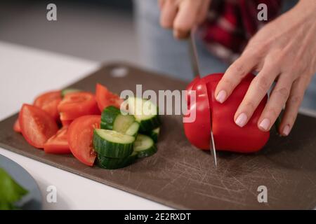 Cutting in slices a fresh vegetable salad young housewife cut sweet pepper, cucumber, tomato on board preparing for a family dinner standing in the Stock Photo